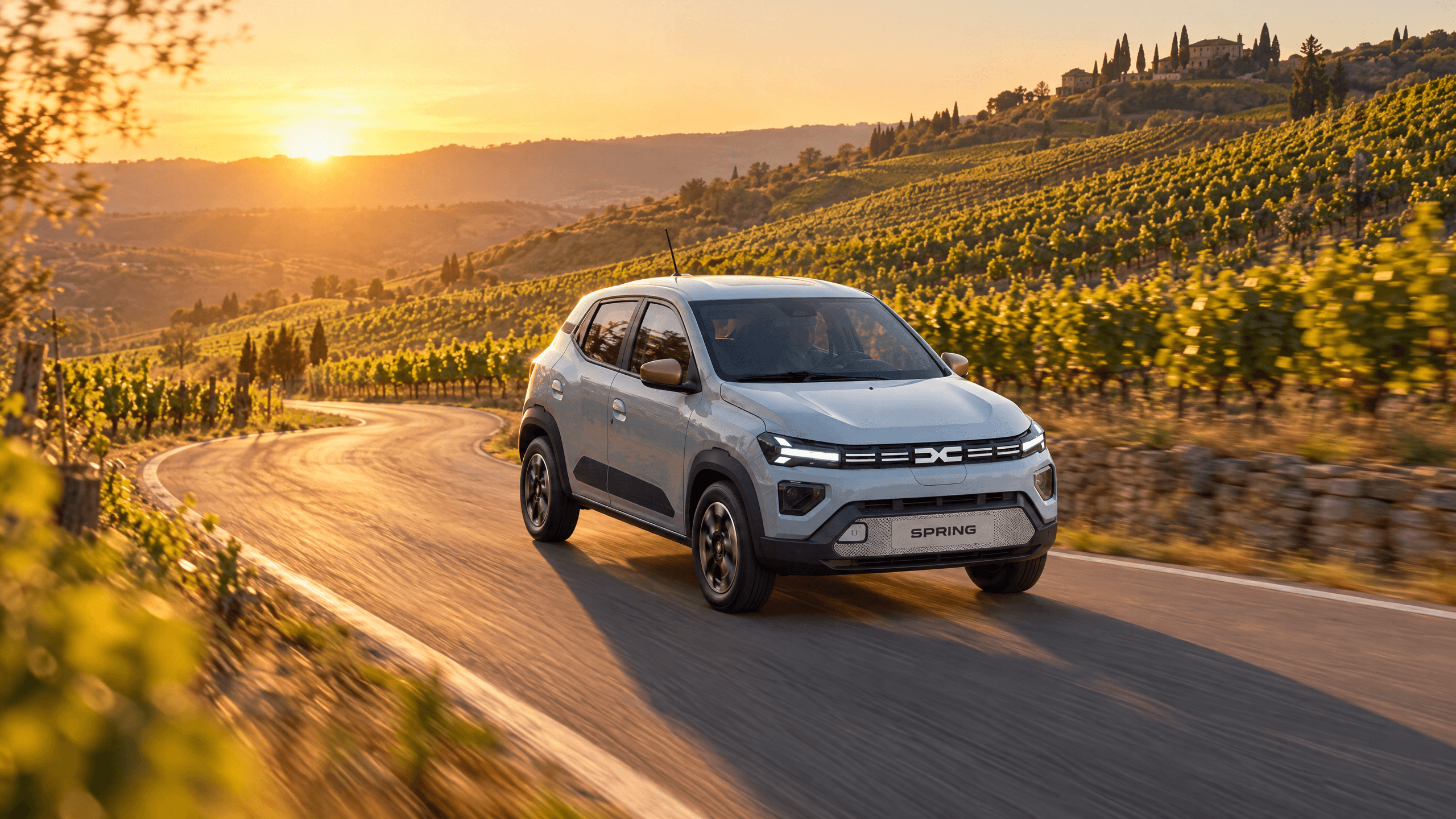 Dacia Spring pe un drum cu serpentine printre dealuri cu vii, lumină de apus